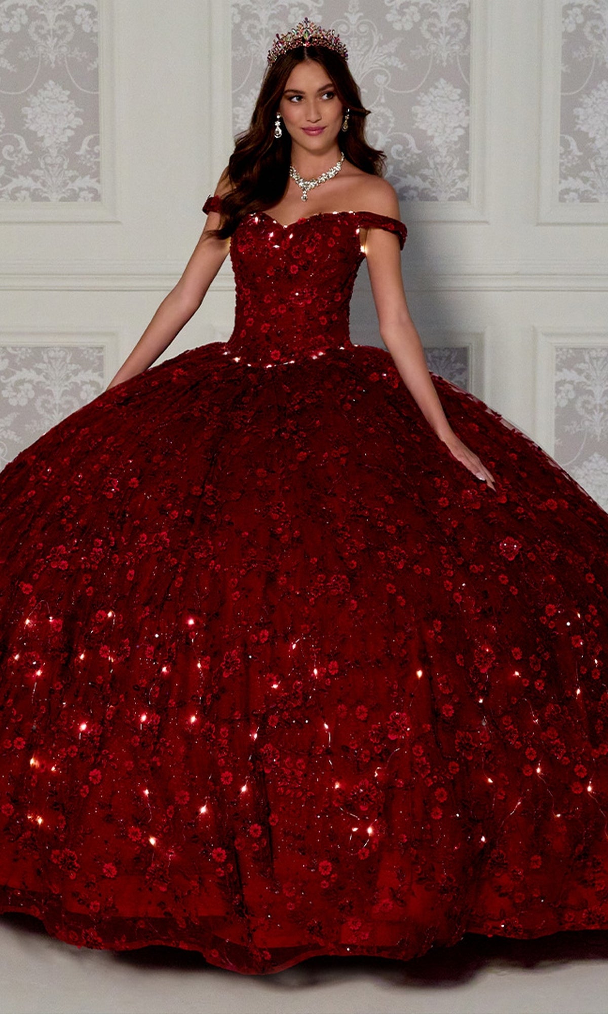 A woman in the Princesa Glow-in-the-Dark Quince Dress PR12266, featuring a red off-the-shoulder, floor-length design with floral details and a tiara, stands before an elegant white background.
